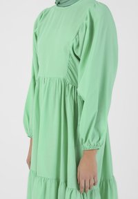 Vihreä, pitkähihainen mekko, joka on valmistettu kevyestä kankaasta. Siinä on rypistetty pääntie, kerrosmainen hame ja elastiset hihansuut. Sujuva pinta.