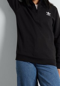 Svart Adidas-hoodie med kvartdragkedja, ribbade muddar och fåll. Liten vit logotyp på bröstet. Blå denimjeans synliga.