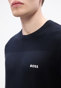 Gros plan d’une personne portant un pull en maille bleu marine avec « BOSS » imprimé en blanc sur la poitrine.