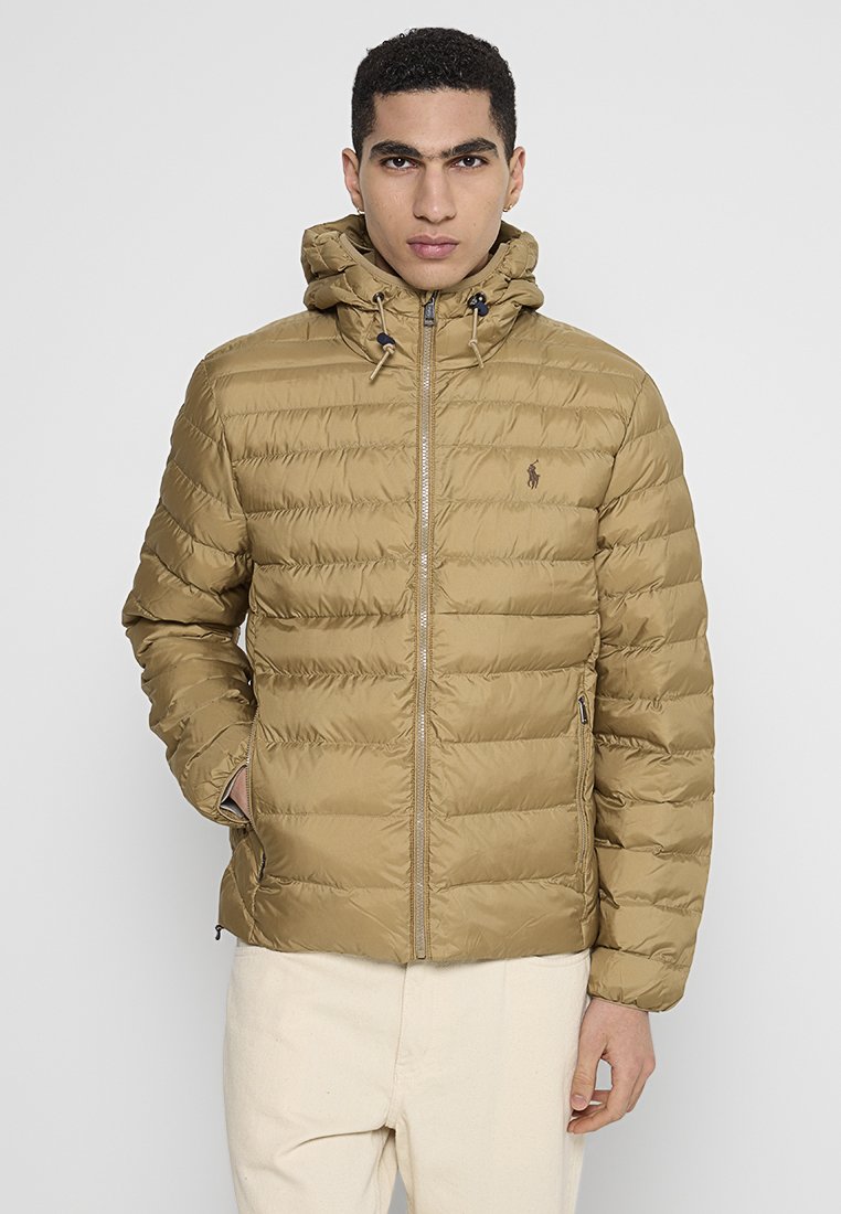 Hellbraune Pufferjacke mit horizontalen Nähten, einem Reißverschluss vorne und einer Kapuze. Sie hatseitliche Taschen und ein kleines Logo auf der linken Brust.