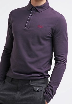 Mann trägt ein eng anliegendes, dunkelviolettes Langarm-Poloshirt mit drei schwarzen Knöpfen und dunkelgraue Hose mit geknöpfter Tasche.