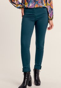 Pantaloni skinny in denim teal con vita alta e orli arrotolati, abbinati a stivali alla caviglia neri. Texture liscia, design aderente.