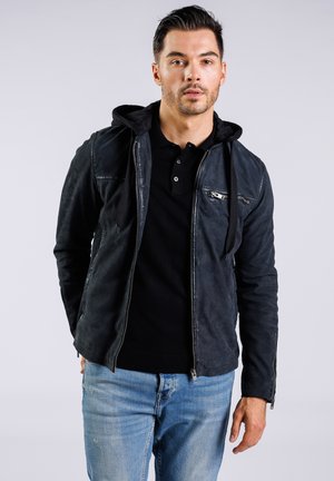Chaqueta de cuero negra con capucha, cierre delantero y bolsillos laterales. Acabado texturizado, estilo ajustado, usada sobre una camiseta polo negra y jeans.