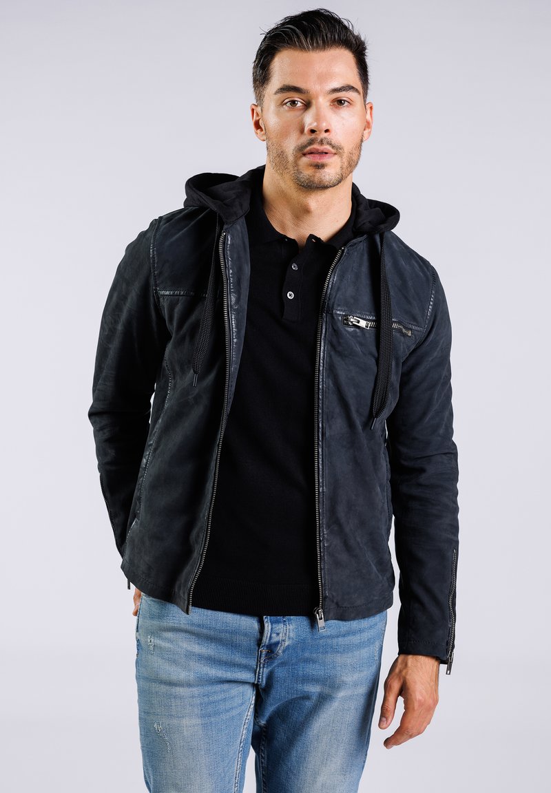 Svart läderjacka med huva, dragkedja framtill och sidofickor. Texturerad yta, figursydd stil, bärs över en svart polo skjorta och denim.