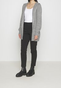 Jasnoszary dzianinowy kardigan z kapturem, długimi rękawami i naszywanymi kieszeniami, noszony na białej koszulce na ramiączkach i czarnych dopasowanych spodniach.