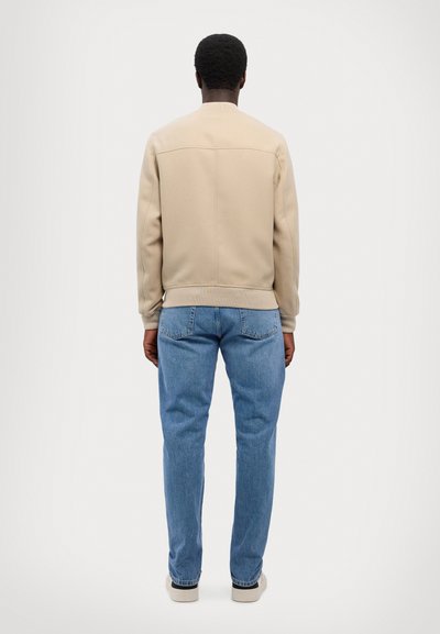 Beige bomber-tagi sileva tekstuuriga, soonikkoes varrukate ja äärisega, kokku sobitatud helesiniste sirgete pükstega ja valgete tossudega.