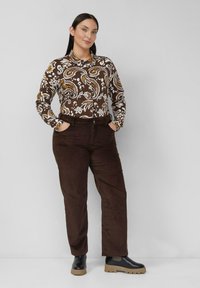 Femme aux cheveux foncés portant une chemise à manches longues à motifs marron et blanc, un pantalon marron foncé et des bottes noires, debout les mains dans les poches.