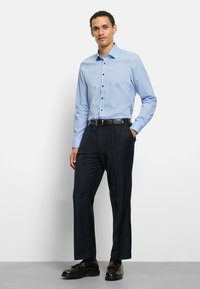 Hellblau kariertes Hemd mit einem Button-Down-Kragen, kombiniert mit dunkelblauen Hosen und braunen Schuhen. Das Hemd verfügt über dezente kontrastierende Musterdetails.