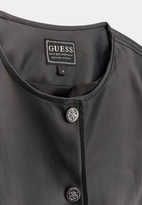 Svart konstläderplagg med rund halsringning, som har två dekorativa silverknappar och en "GUESS"-etikett inuti. Storleksmärke synligt.