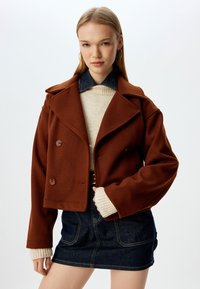 Giacca corta marrone con design doppiopetto, ampio colletto e dettagli con bottoni, abbinata a un maglione beige e una gonna di jeans.