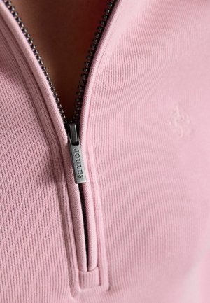 Nærbillede af en delvist lynlåst lyserød ribstrikket sweater med en synlig metallynlås trække, der er indgraveret med "Joules."