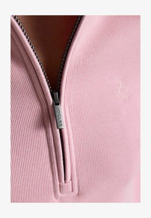 Prim-plan al unui pulover roz cu dungi, parțial fermecat, cu un mâner metalic al fermoarului vizibil, gravat cu "Joules".