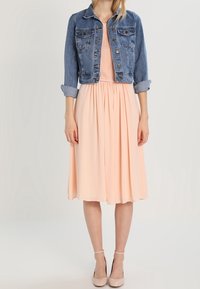 Veste en denim bleue, à manches courtes, avec un devant à boutons. Robe pêche avec un corsage plissé, longueur genou, en tissu fluide. Talons nude.