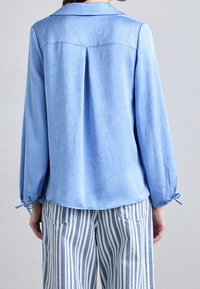 Světle modrá, texturovaná blůza s límcem a dlouhými rukávy, s nařasenými manžetami. V kombinaci s horizontálně pruhovanými kalhotami ve světle modré a bílé barvě.