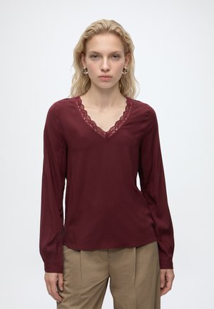 Jeune femme aux cheveux blonds ondulés portant un chemisier bordeaux à manches longues avec un col en dentelle et un pantalon beige, debout devant un fond uni.