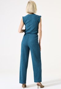 Teal jumpsuit met een gestructureerde afwerking, mouwloos topje, ruches op de schouders en wijde broekspijpen. Draag het met sleehakken.