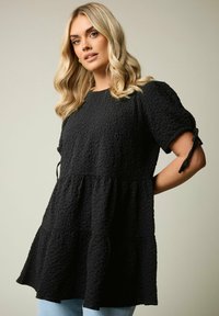 Top nero con texture, caratterizzato da una vestibilità comoda, maniche corte con lacci e un design a strati. Il tessuto morbido accentua la profondità visiva.