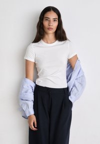 T-shirt bianca aderente, con scollo rotondo e maniche corte, abbinata a una camicia a righe blu chiaro, aperta e con bottoni, e pantaloni scuri su misura.