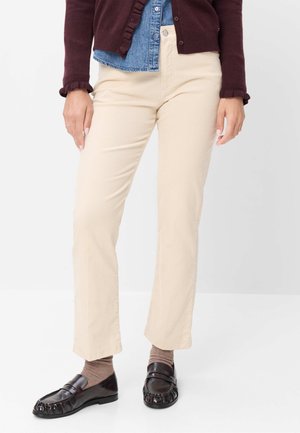 Person iført beige bukser, brune sokker, sorte loafers, en cowboyskjorte og en mørkebrun cardigan mod en hvid baggrund.