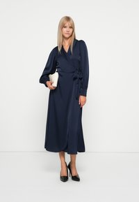 Navy wrap dress in smooth fabric with long sleeves, a tie at the waist, and a mid-length hem. Worn with black high heels and a small white clutch.