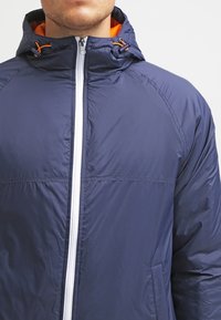 Giacca blu navy in materiale leggero, con cerniera bianca e dettagli arancioni, dotata di cappuccio e due tasche laterali. Tessuto dalla texture liscia.