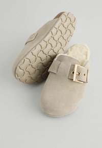 Beige mockasklackar med en mjuk krämfärgad foder, en guldbucklad detalj och en texturerad gummisula med ett geometriskt mönster.