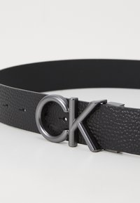 Schwarzer Ledergürtel mit strukturiertem Finish und einer silbernen Schnalle mit stilisiertem "CK"-Logo, verstellbar mit mehreren Löchern.