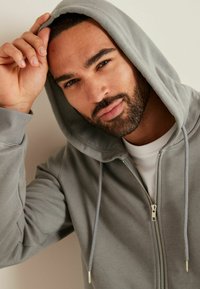 Helle graue Zip-Hoodie mit Kapuze zum Verstellen, strukturiertem Stoff und silbernem Reißverschluss. Wird über einem weißen T-Shirt getragen, Modell stellt die Kapuze ein.
