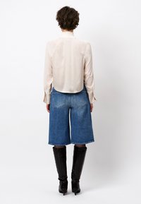 Beige satijnen blouse met ronde zoom, gecombineerd met blauwe denim culottes. Het model draagt kniehoge zwarte laarzen met hakken en een gestructureerde afwerking.