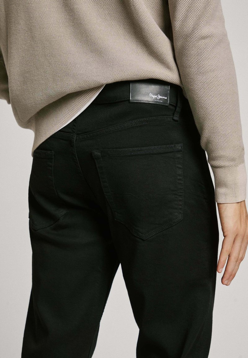 Schwarze Denim-Hosen verfügen über Gesäßtaschen und ein Lederlabel am Bund. Ein lockerer beiger Strickpullover wird über die Hosen getragen.
