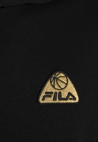 Tela negra con un logo bordado en dorado, texturizado, que presenta una pelota de baloncesto y la palabra "FILA" en forma triangular.