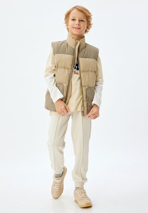 Gilet imbottito beige con colletto alto, indossato sopra una camicia a maniche lunghe color crema. Abbinato a joggers chiari e sneakers beige.