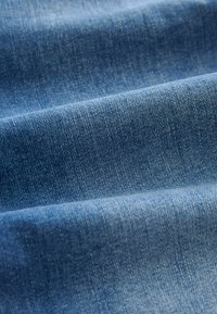 Denim tkanina u srednje plavoj nijansi, s glatkom teksturom i suptilnim twill tkanjem. Površina pokazuje blage varijacije.