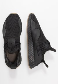Svarta mesh-sneakers med en texturerad yta, rund tå och en brun gummisula. Har svarta snörningar och en logotyp på tungan.