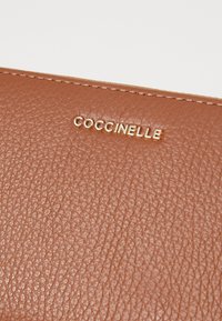 Portefeuille en cuir brun avec une finition texturée, arborant un logo doré "Coccinelle". Des détails cousus mettent en valeur les bords.