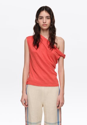 Mujer con cabello largo y oscuro que lleva una camiseta roja sin mangas con un hombro descubierto y pantalones crema con rayas de colores en los lados.