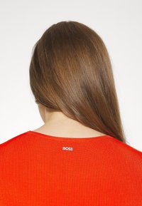 Personne aux longs cheveux bruns portant un haut rouge côtelé avec un petit logo "BOSS" en haut du dos, sur un fond blanc uni.