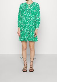 Vestido floral verde con mangas largas y un escote con lazo. Presenta una falda de volantes que muestra patrones de hojas blancas. Combinado con sandalias negras con cordones.