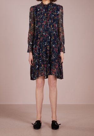 Marineblauwe gebloemde jurk met lange mouwen, met geruite kraag en zoom, gedragen met zwarte platte loafers met decoratieve gespen.