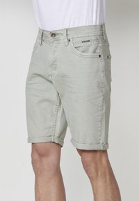 Vyras, vilkintis šviesiai pilkas džinsines šortus su sukamuoju apvadu ir baltą marškinį, stovi prieš paprastą šviesią foną.