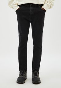 Jean en denim noir coupe slim, avec fermeture par bouton, poches avant et légère usure près des genoux. Porté avec des chaussures foncées.