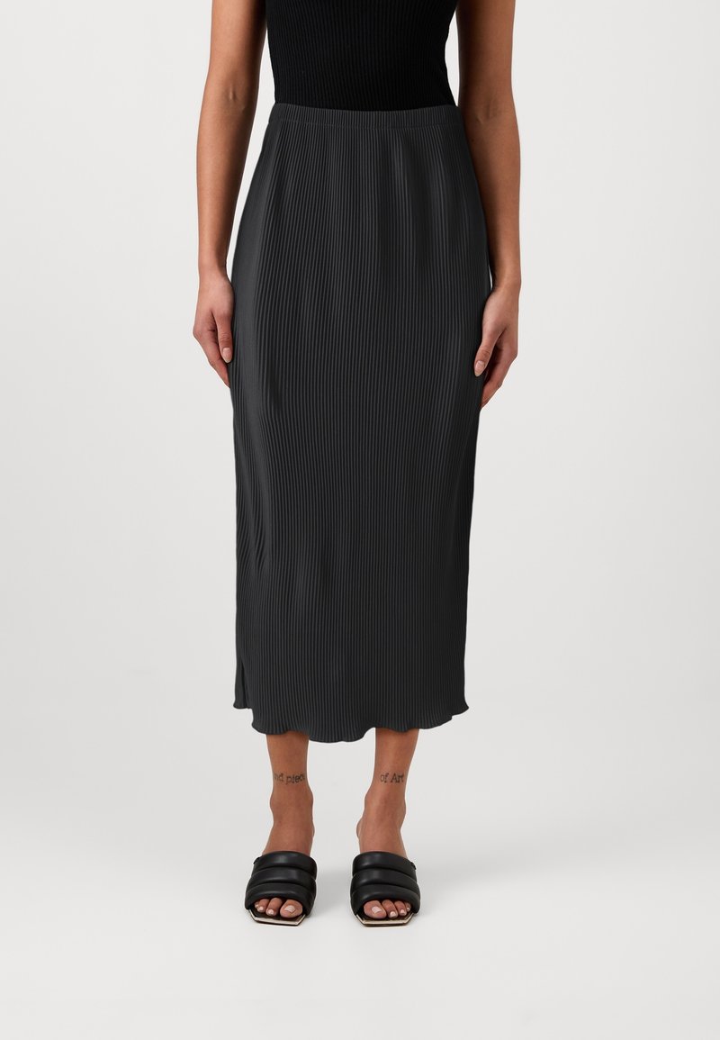 Femme portant une jupe midi côtelée noire, un haut sans manches noir et des sandales à enfiler noires rembourrées, debout devant un fond uni.
