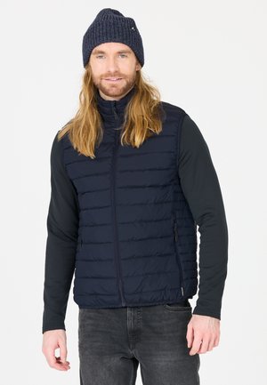 Marineblauwe puffer vest met een verticale gestikte patroon, rits aan de voorkant, hoge kraag en zijzakken; gedragen over een zwart langarmshirt.