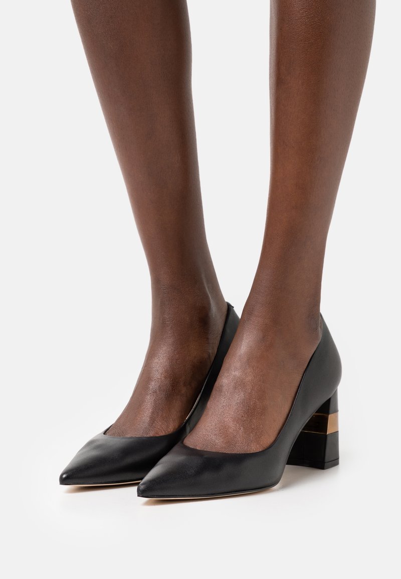 Escarpins en cuir noir à bout pointu avec talons carrés ornés d'une bande dorée, portés sur des pieds à la peau foncée sur un fond uni.