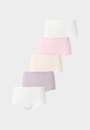 Femme aux longs cheveux foncés portant un soutien-gorge en dentelle blanche et un sous-vêtement blanc à motifs, debout devant un fond clair uni.