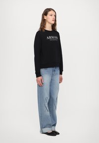 Zwarte sweatshirt met het "ARMANI EXCHANGE" logo, gedragen met lichtblauwe loszittende jeans met een subtiele patroonprint op de benen. Zwarte flats.
