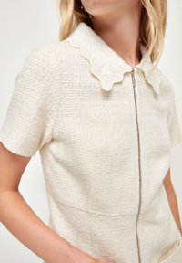Chaqueta blanca texturizada con mangas cortas, un cuello festoneado y cremallera al frente. El material parece tejido con un brillo sutil.