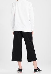 Vit sweatshirt med långa ärmar, svarta korta byxor och svarta sneakers med vita detaljer. Bakgrunden är en enfärgad yta.