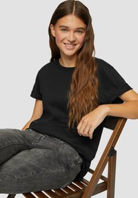 Zwarte katoenen t-shirt met een relaxte pasvorm, korte opgerolde mouwen en een ronde hals, gecombineerd met grijze spijkerbroek, zittend op een houten stoel.