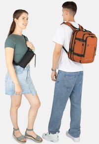 Schwarze Umhängetasche mit Kordelzug, getragen von einer Frau in grünem Oberteil und Jeansrock; Mann in weißem T-Shirt und orangefarbenem Rucksack, blaue Jeans.